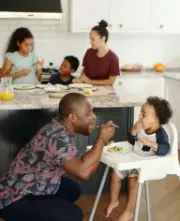 Father feeds toddler in a highchair, family eating at kitchen island.