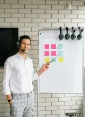 Man with headset points at a whiteboard with sticky notes in front of a brick wall.