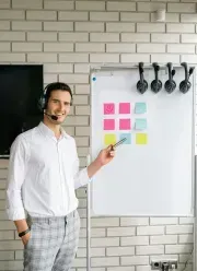 Man with headset points at a whiteboard with sticky notes in front of a brick wall.