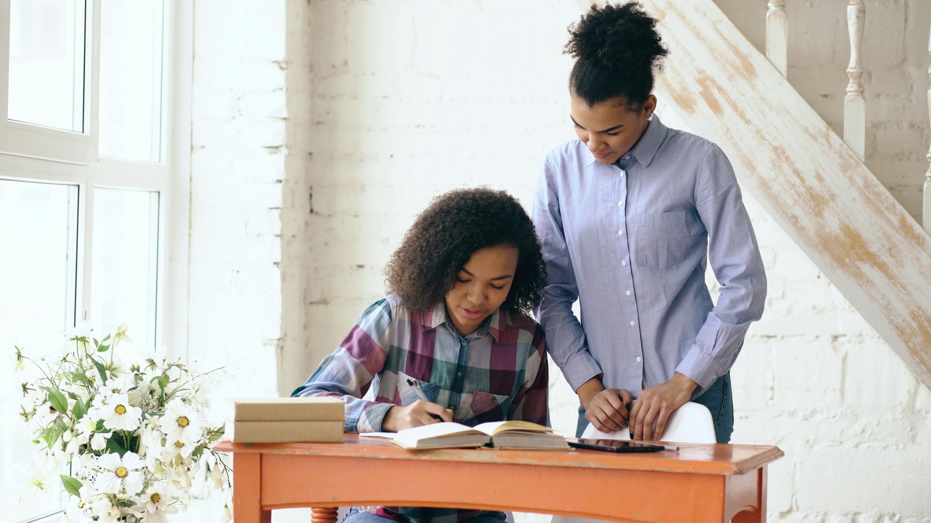 Woman with glasses assisting a child drawing with colored pencils at a table.