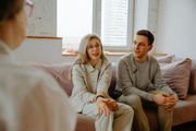Couple on a couch talking to a person, likely a therapist, in a light-filled room.