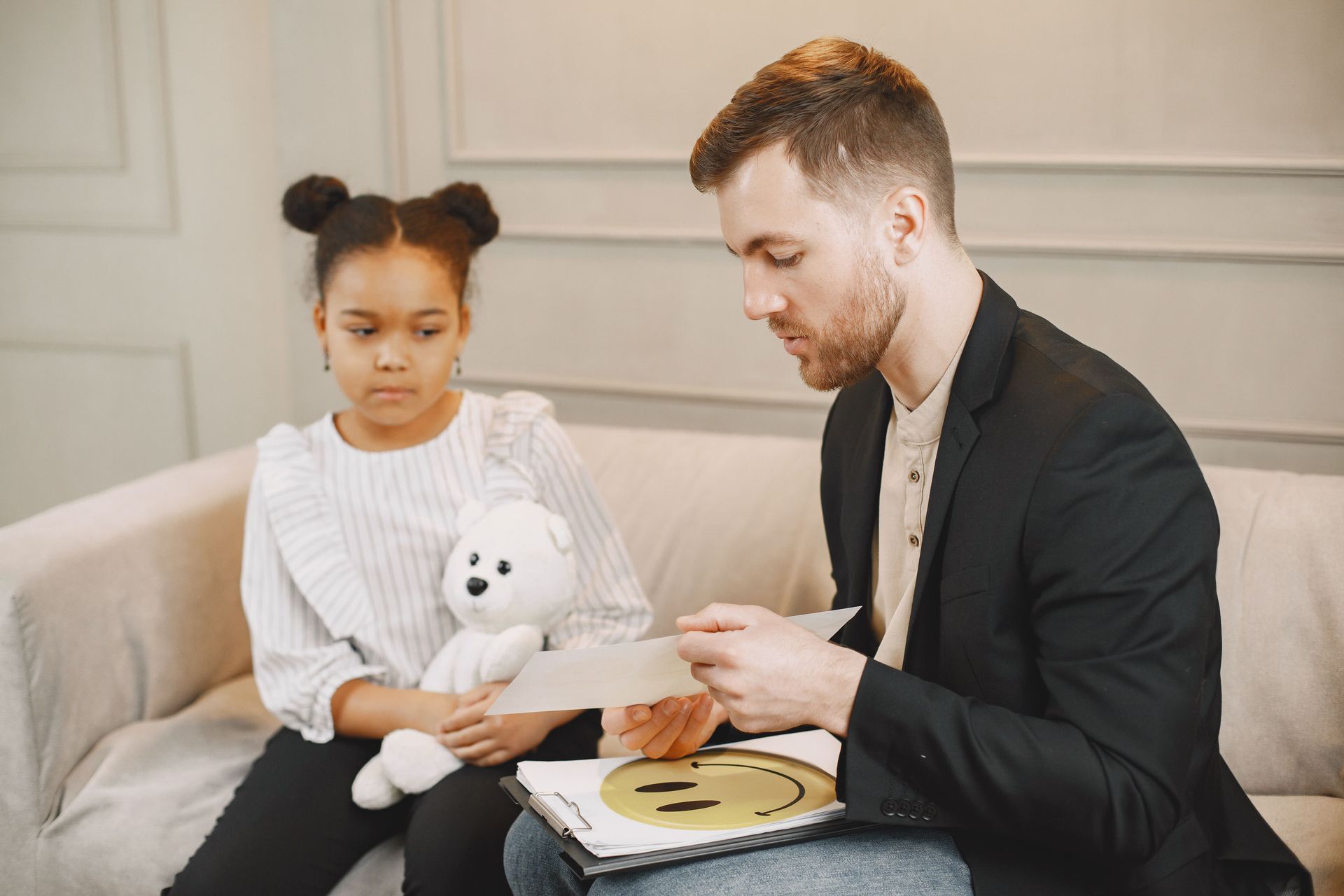 Woman showing flashcards to a child, seated on a colorful mat. An easel and sofa are in the background.