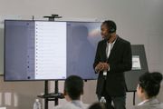 Man in suit presents to a group, with a large screen displaying text and a camera above.