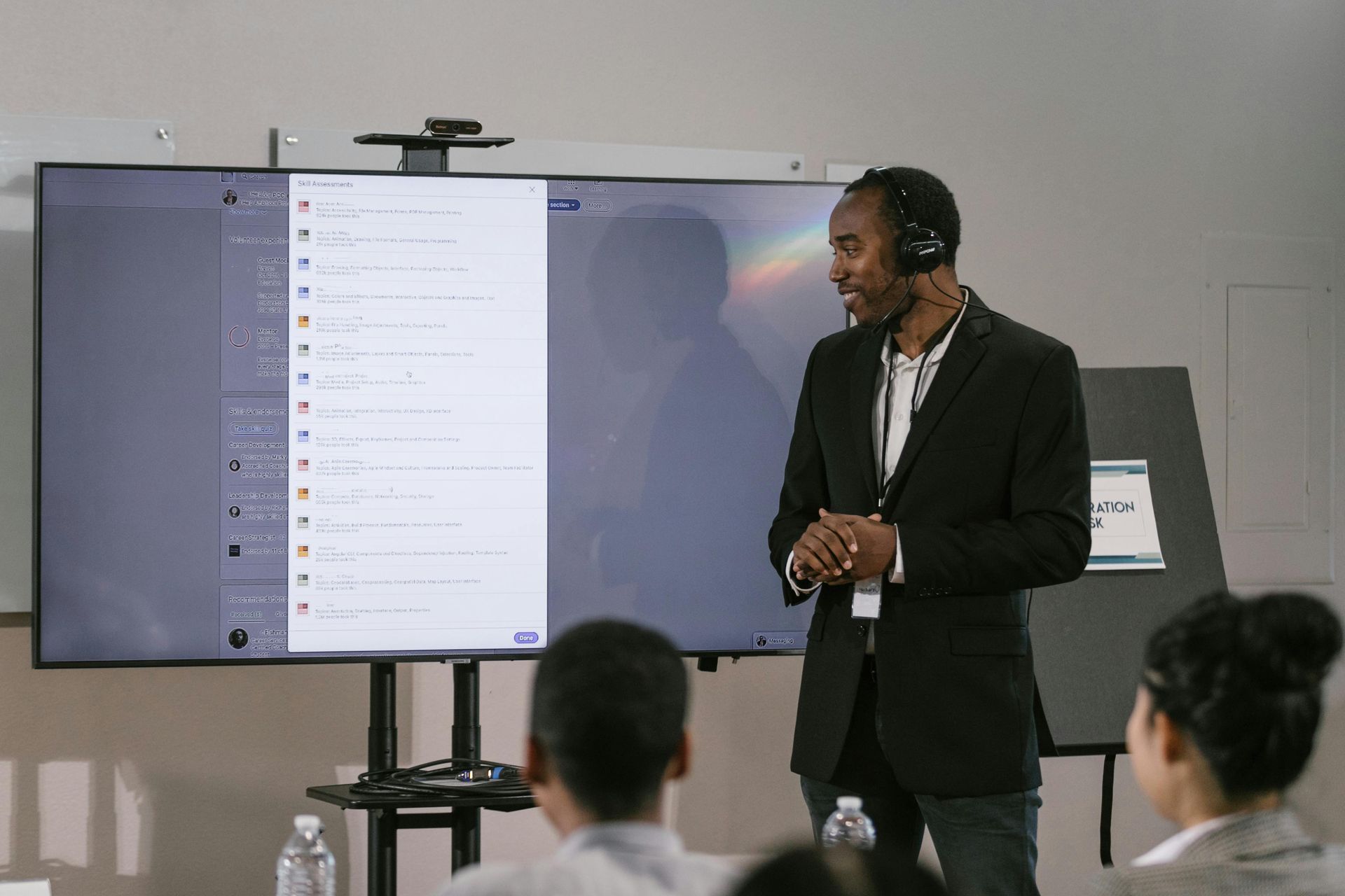 Man in suit presents to a group, with a large screen displaying text and a camera above.