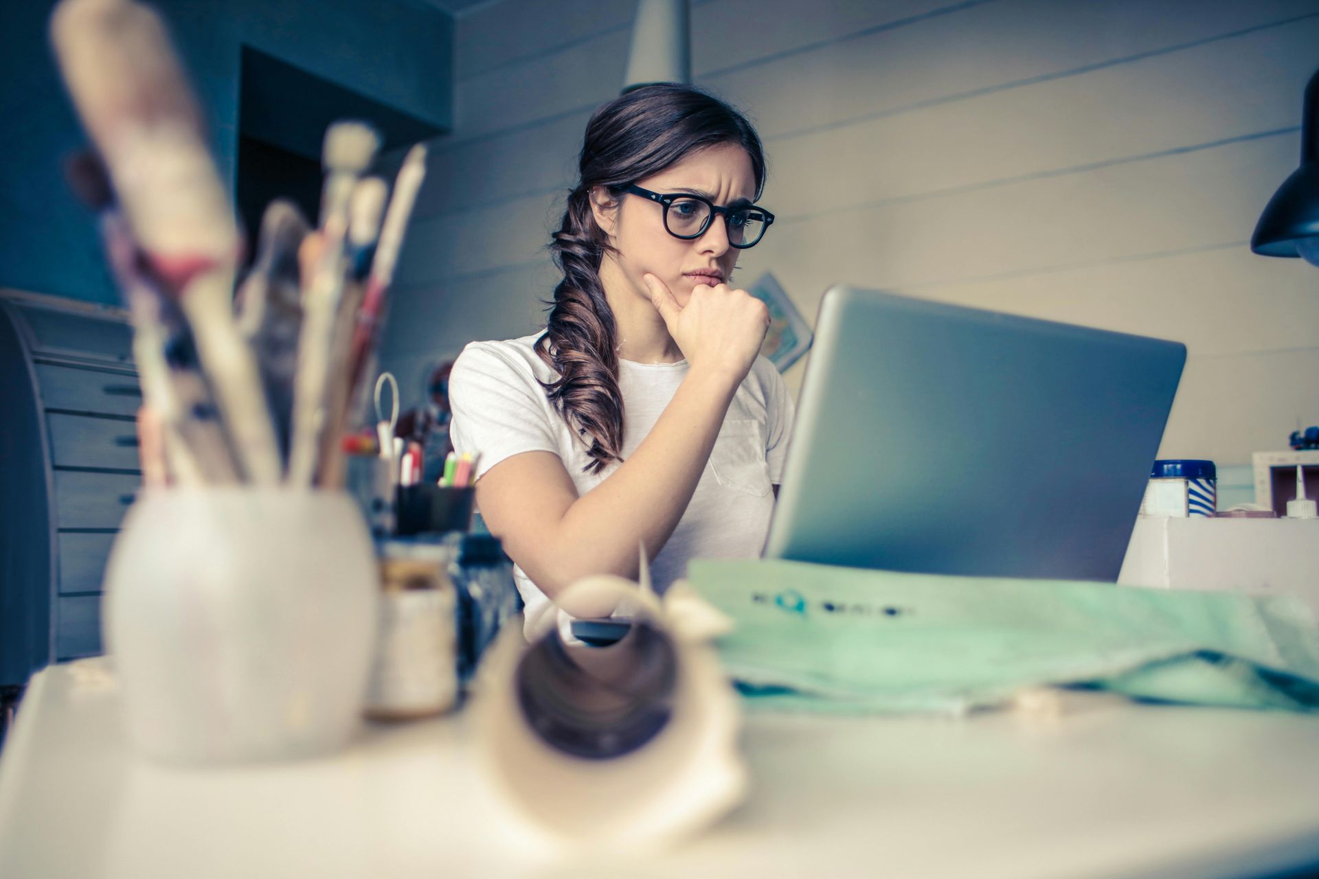 Woman with glasses looks intently at a laptop, hand on chin, paintbrushes in a vase on the table.