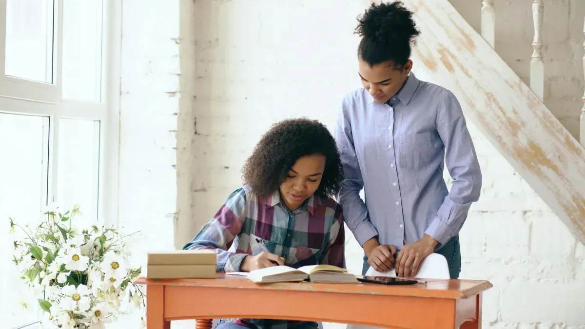 Woman with glasses assisting a child drawing with colored pencils at a table.