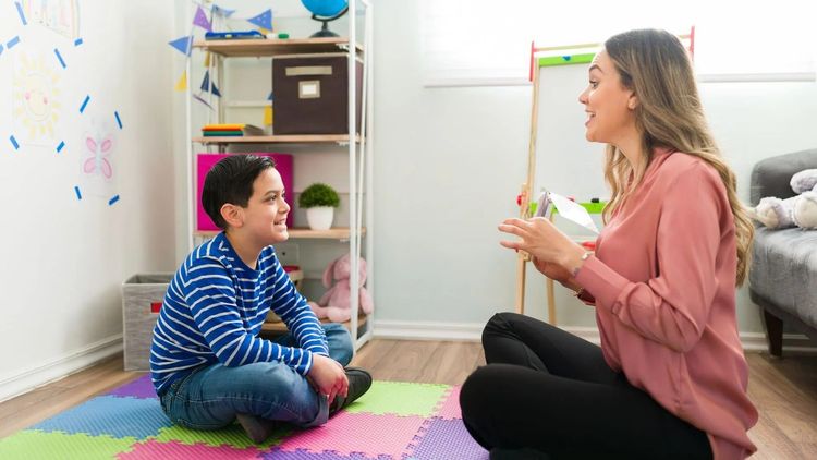 Woman and boy sit on floor, talking in colorful room. Shelves with globe and toys are visible.