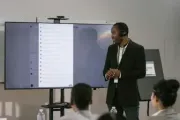 Man in suit presents to a group, with a large screen displaying text and a camera above.