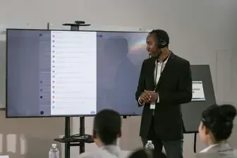 Man in suit presents to a group, with a large screen displaying text and a camera above.