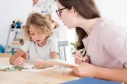 Child drawing at a table with an adult. The adult, wearing glasses, points at paper. Crayons on table.