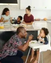 Father feeds toddler in a highchair, family eating at kitchen island.