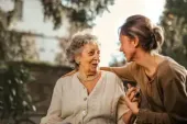 Woman comforting an elderly person outdoors, both smiling, hand on shoulder.