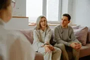 Couple on a couch talking to a person, likely a therapist, in a light-filled room.