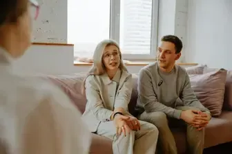 Couple on a couch talking to a person, likely a therapist, in a light-filled room.
