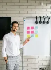 Man with headset points at a whiteboard with sticky notes in front of a brick wall.