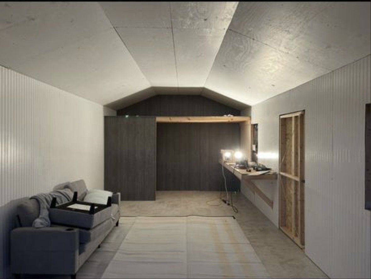 Interior of a room with a gray sofa on the left, white walls, a vaulted ceiling, and a dark wood partition in the back.