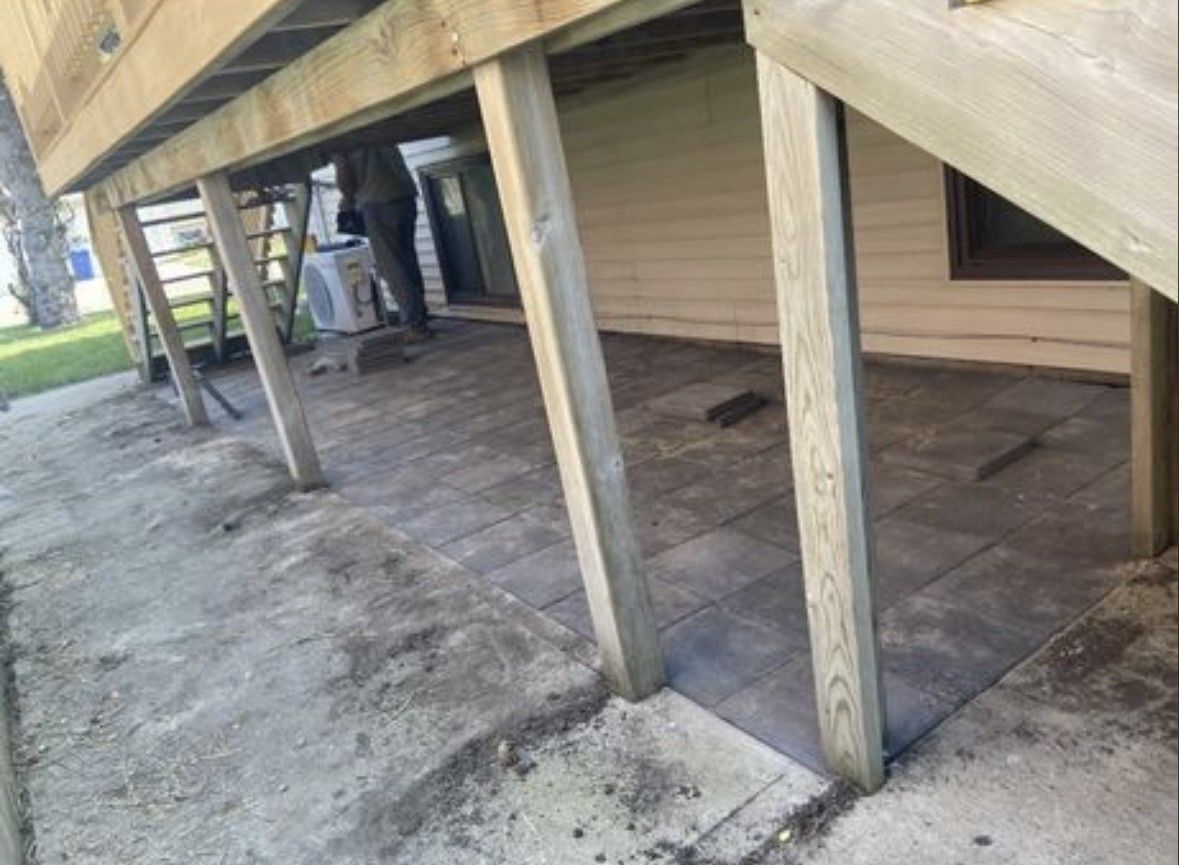A partially finished stone paver patio beneath a wooden deck, with an unpaved dirt area in the foreground.