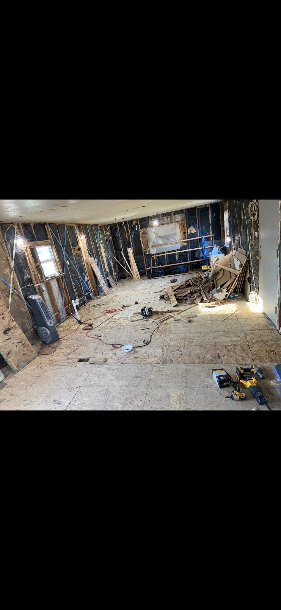 A room undergoing interior renovations with exposed wall studs, subflooring, and debris scattered across the floor.