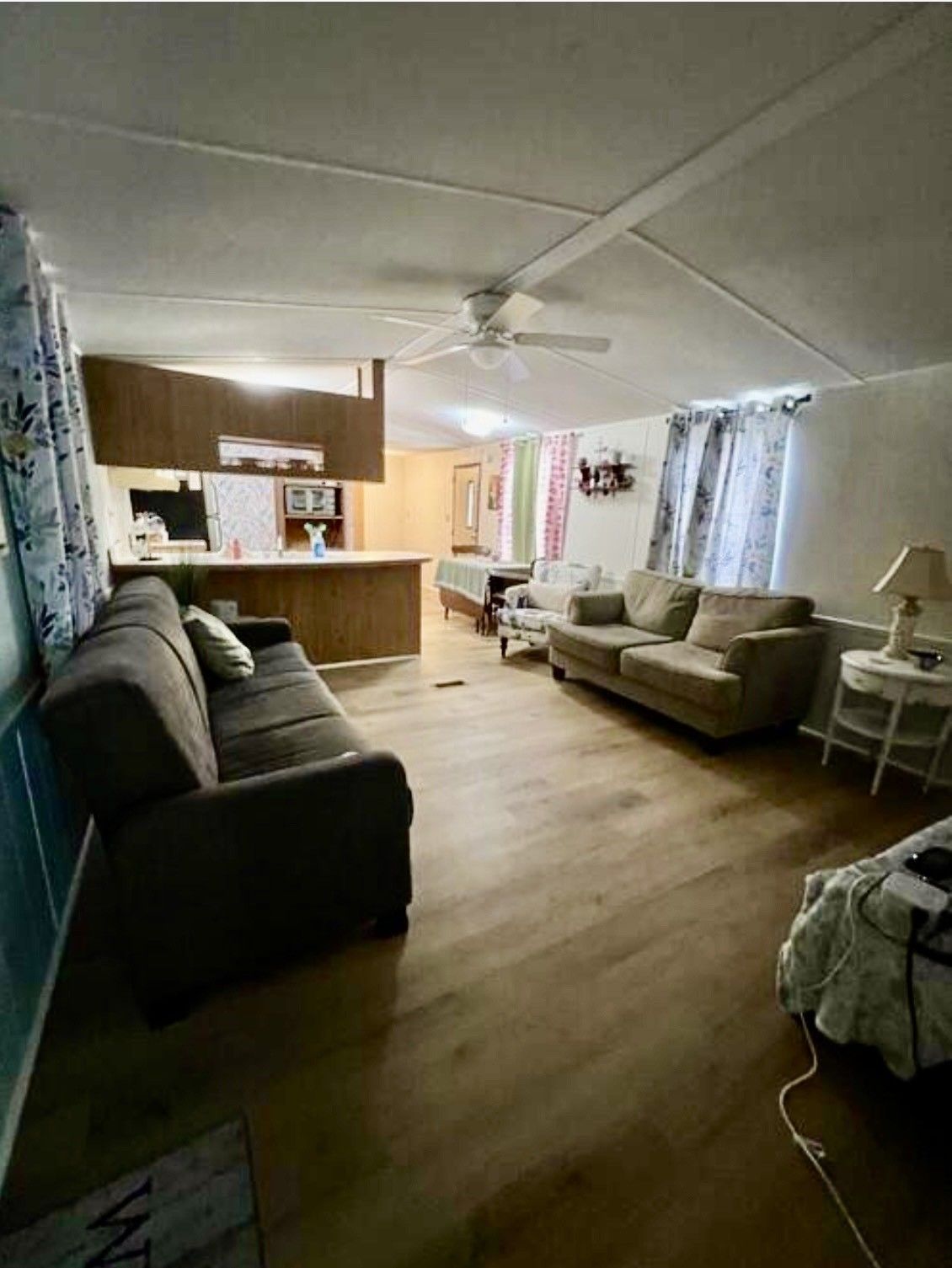 A living room with a dark sofa, a light-colored couch, wood-look flooring, and an open kitchen counter in the background.