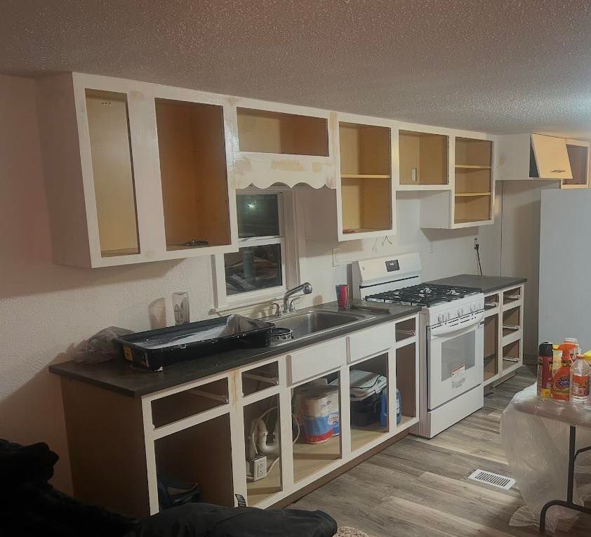 A kitchen undergoing renovation with white cabinets missing their doors, a dark countertop, a sink, and a white stove.