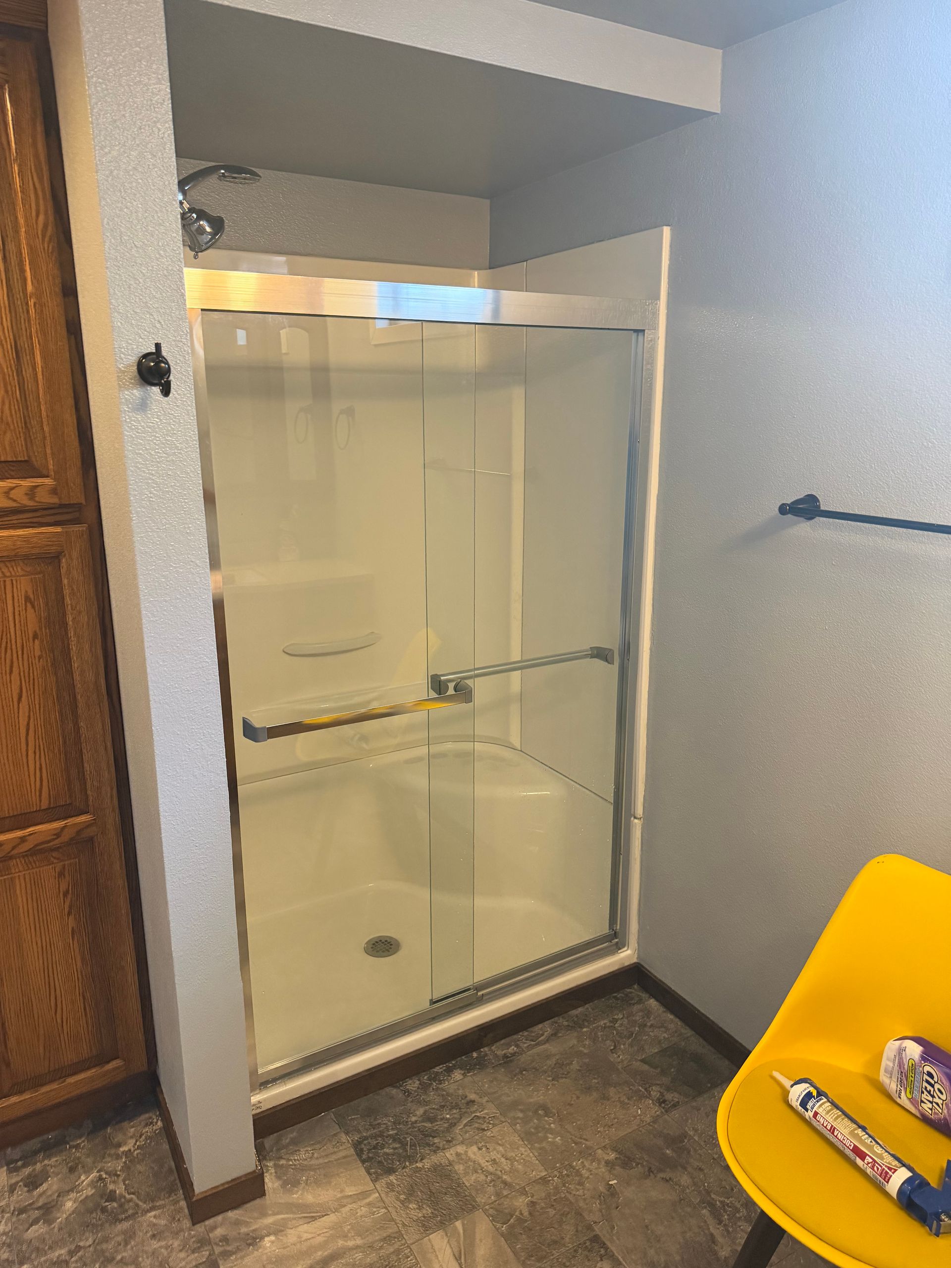 Bathroom shower stall with glass sliding door, beige walls, and a yellow chair in the corner.