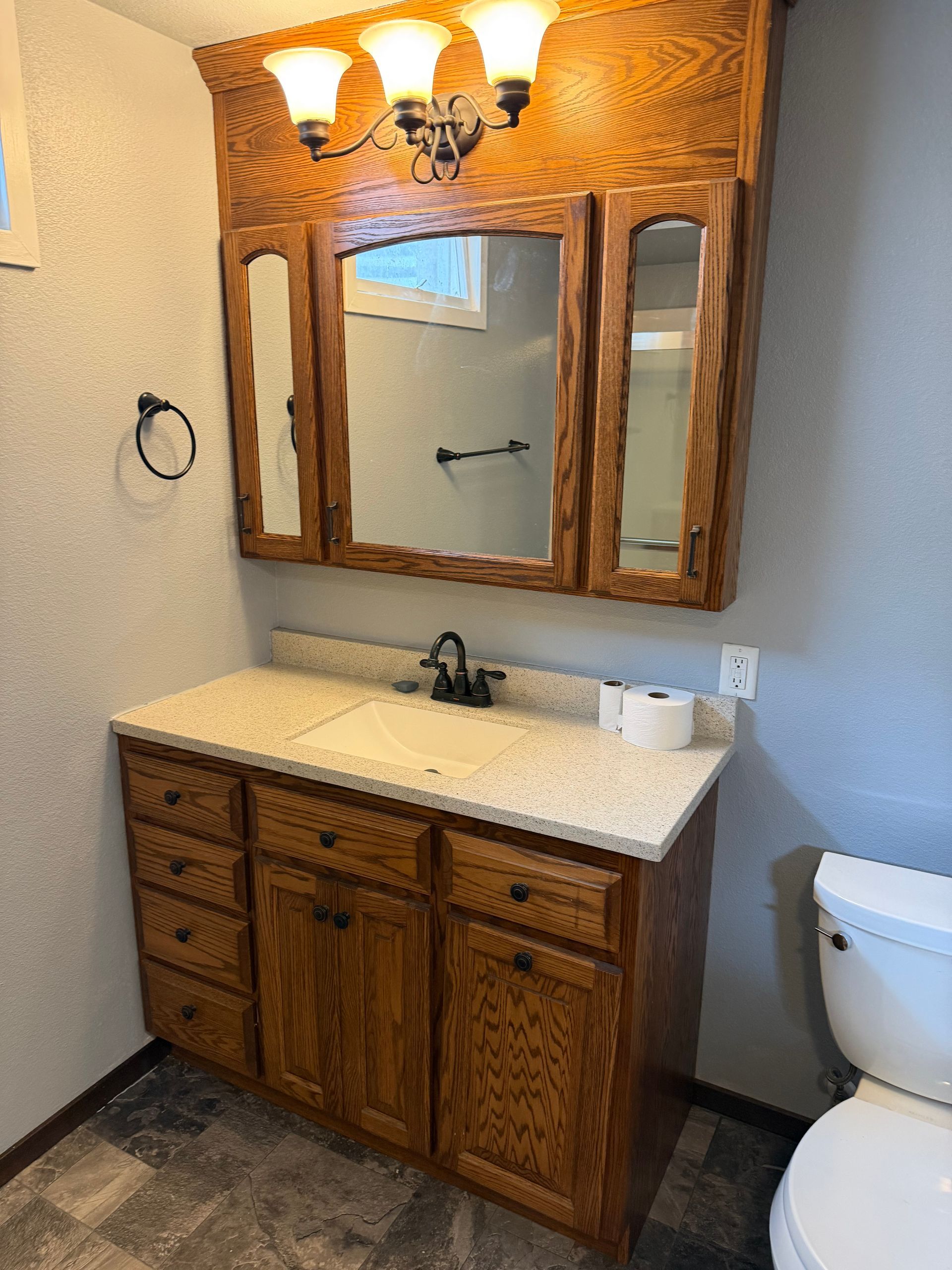 Wood bathroom vanity with mirror, sconce lights, beige countertop, and part of a toilet visible.