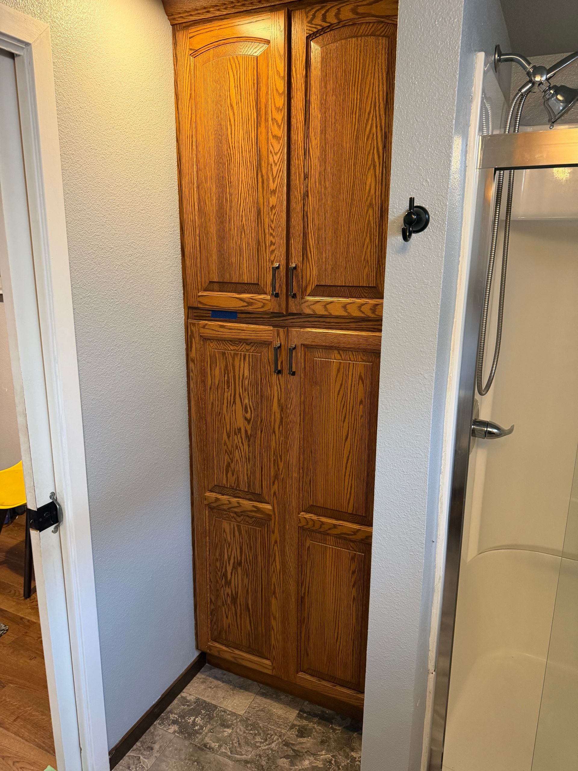 Narrow bathroom closet with wood cabinets between a white wall and shower stall.
