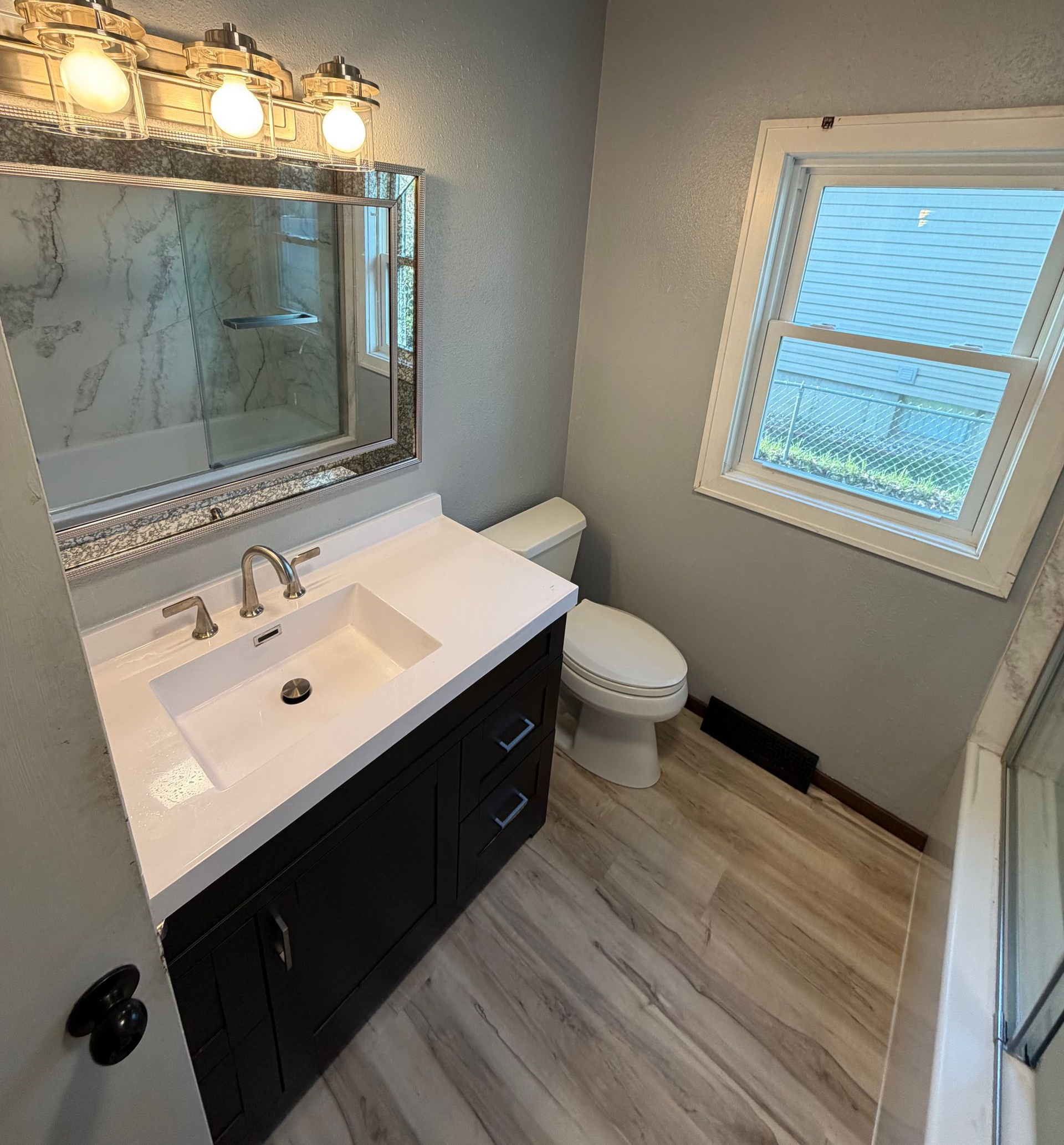 Bright bathroom with white vanity, dark cabinets, ornate mirror, window, and toilet.