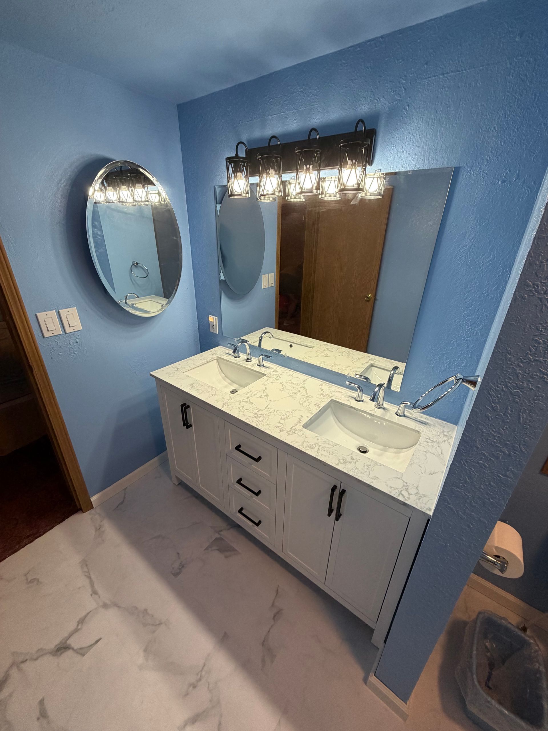 Blue bathroom with double sink vanity, large mirror, oval wall mirror, and towel ring by the door