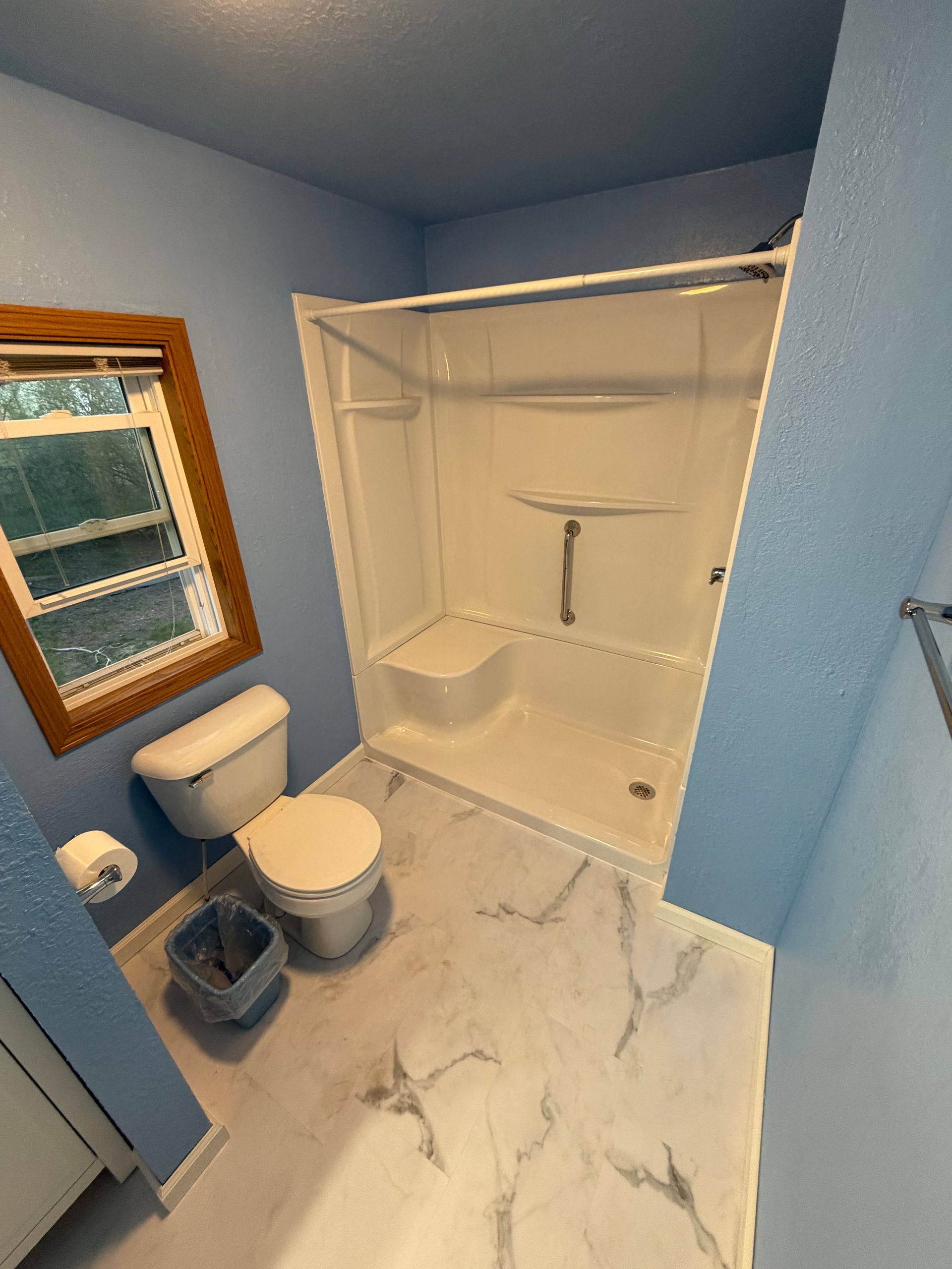 Small blue bathroom with toilet, vanity, window, and white shower-tub combo with curtain rod.