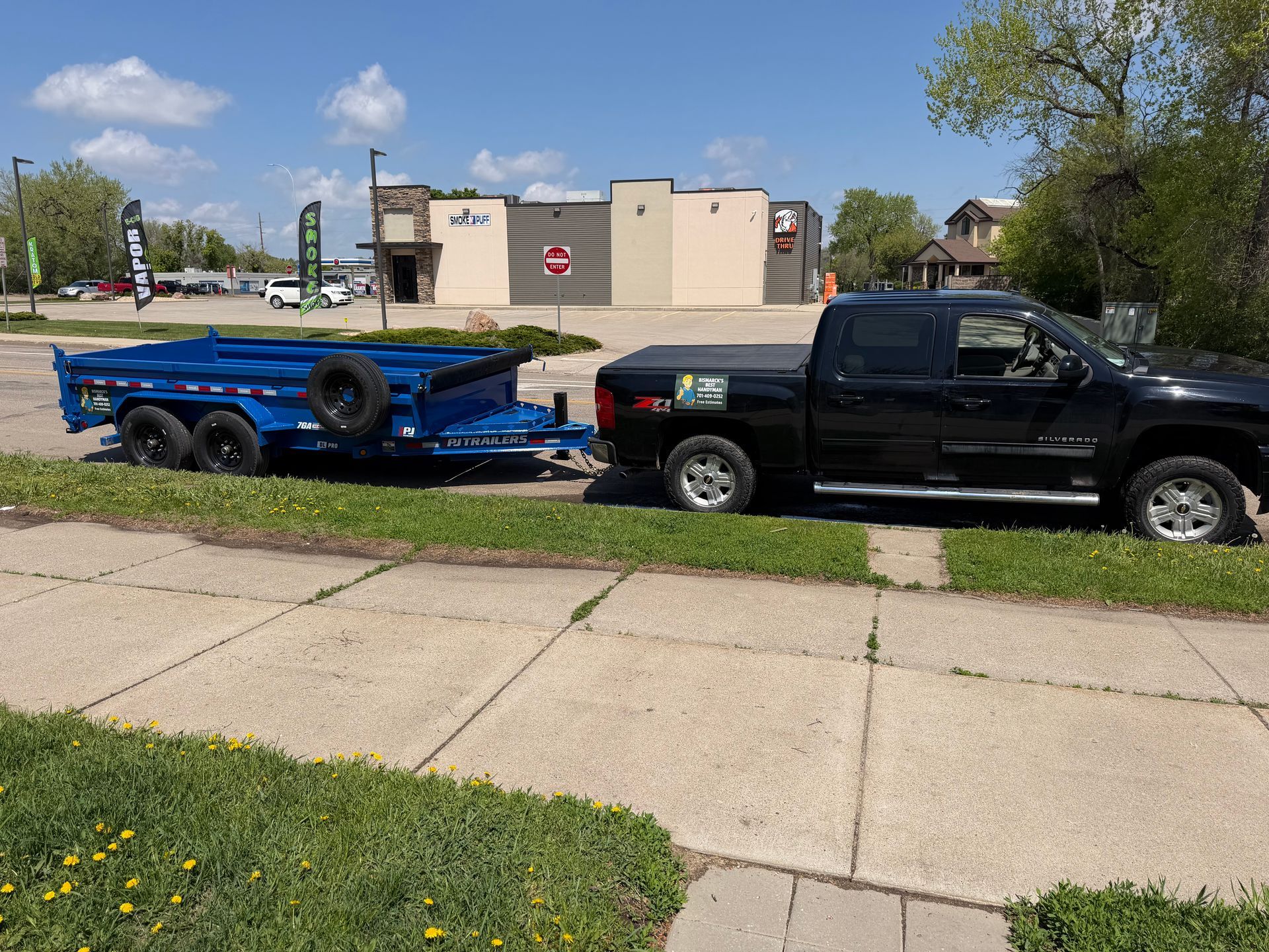 Black pickup truck towing a blue boat trailer parked along a sidewalk near a building.