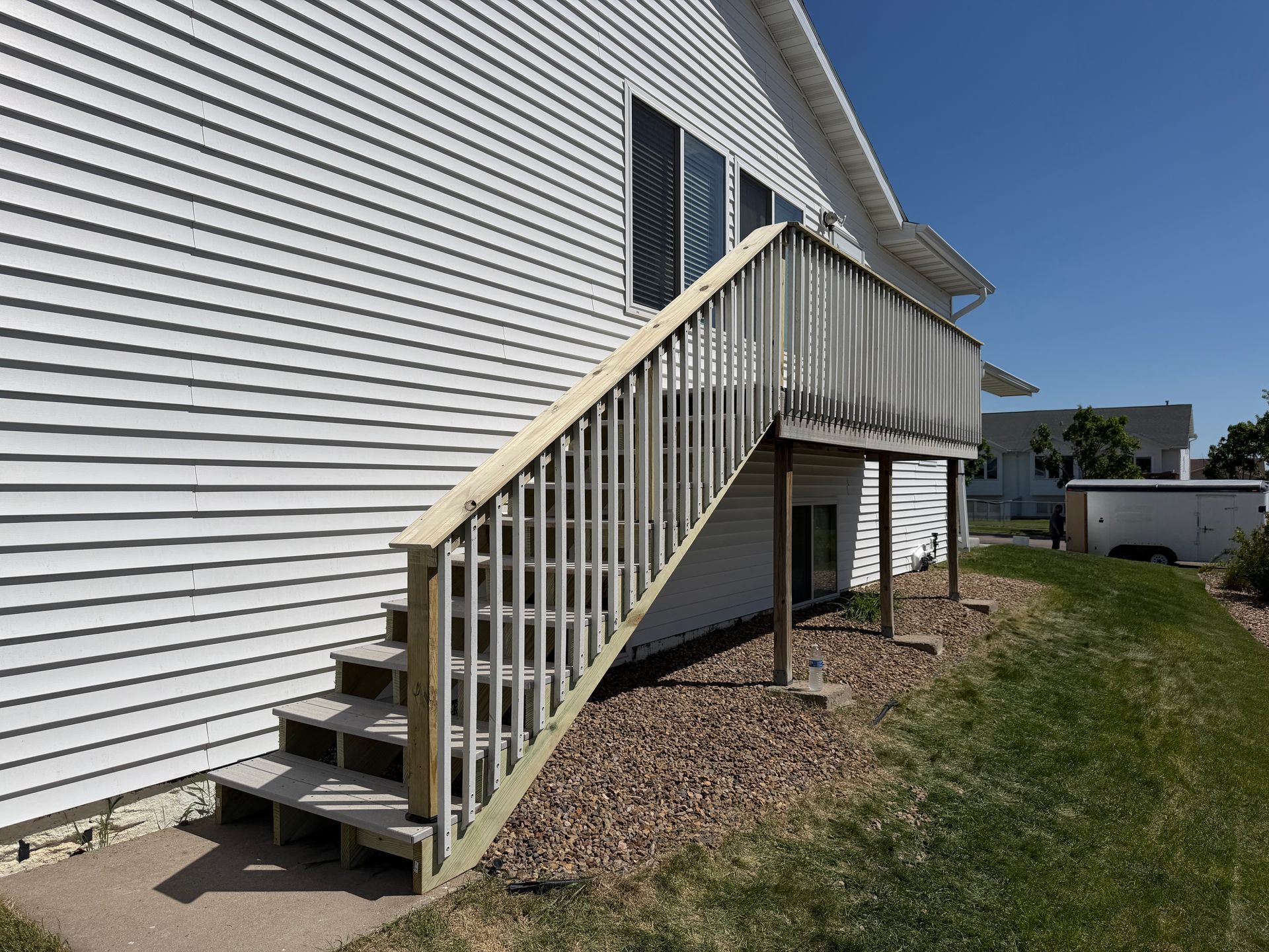 Exterior staircase leading to an elevated deck on a white-sided house, with grass and gravel below