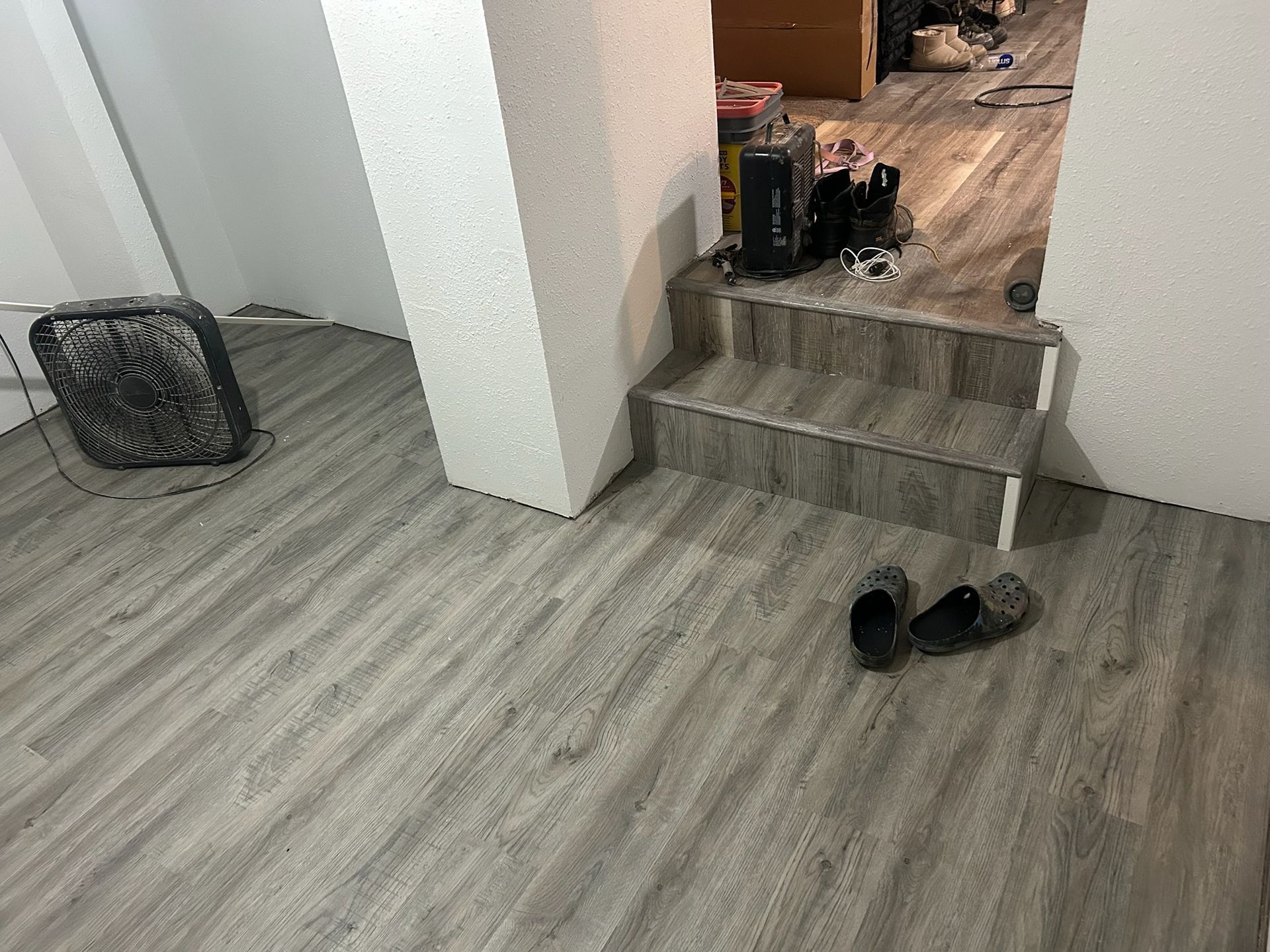 A gray wood-look floor with a box fan, a two-step staircase, and a pair of dark slip-on shoes.
