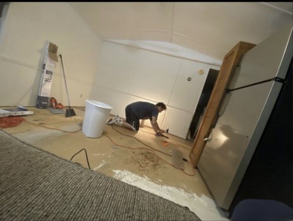 A person kneels on a room's unfinished subfloor, working near a wall. A white bucket and a refrigerator are nearby.