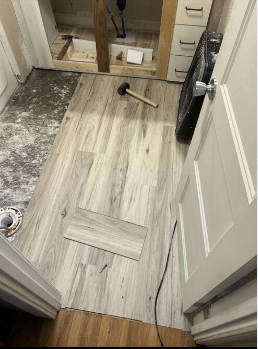 A partially installed light-wood luxury vinyl floor in a bathroom, with a mallet and a loose plank on the floor.