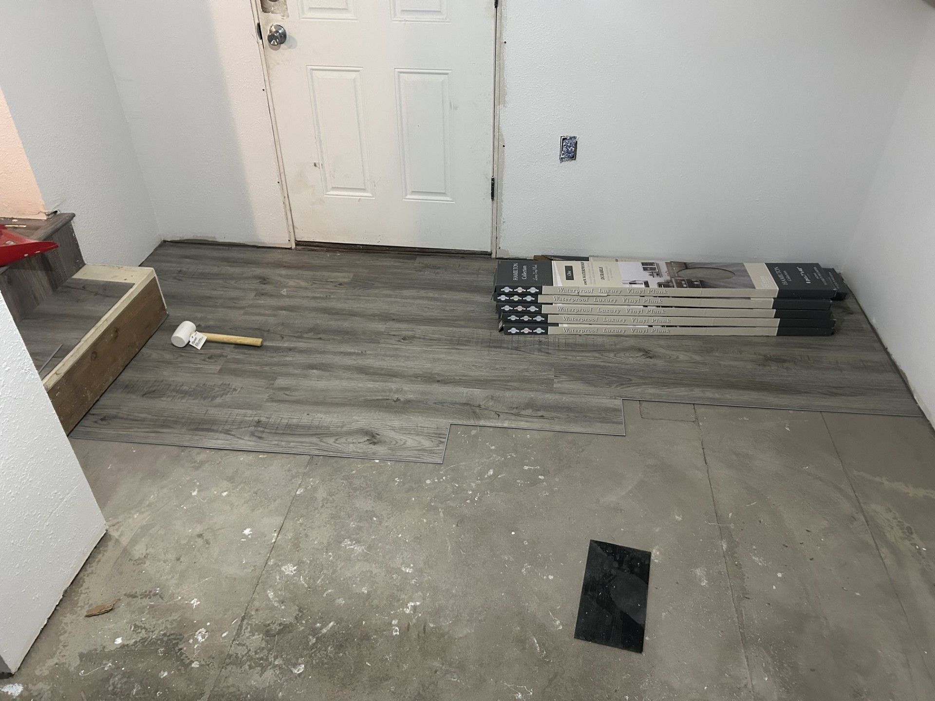 Grey wood-look vinyl planks are partially installed on a concrete floor in a room near a white door and boxes of flooring.