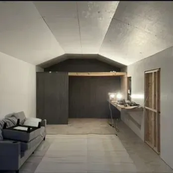 An unfinished room features white walls, a vaulted ceiling, a grey sofa, and a wooden frame for a future doorway.