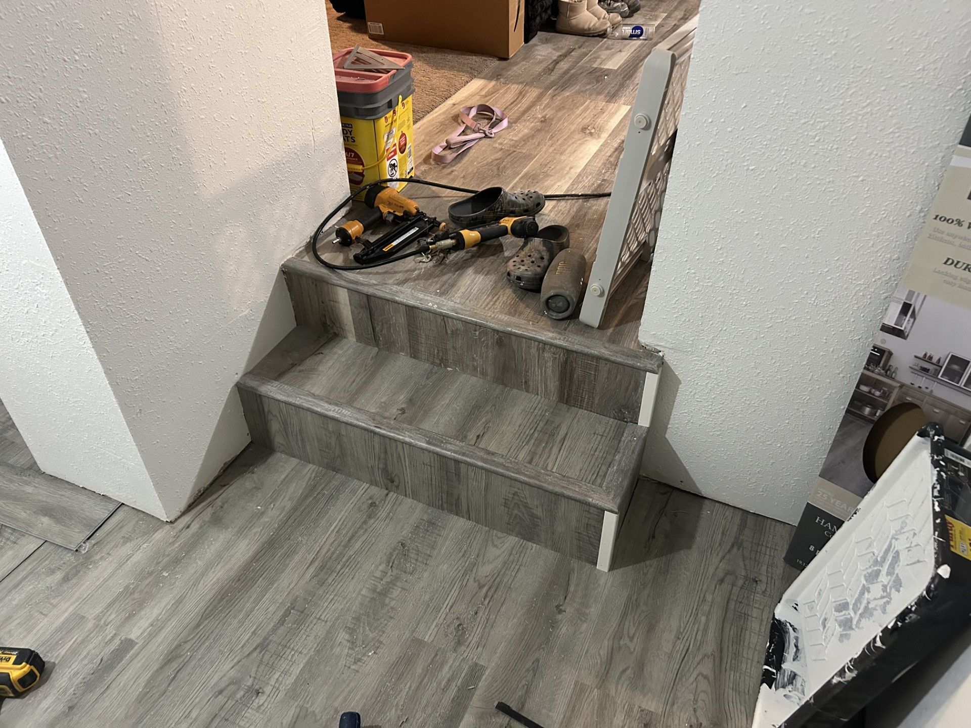 A construction site showing two steps transitioning between rooms with gray wood-look flooring and scattered power tools.