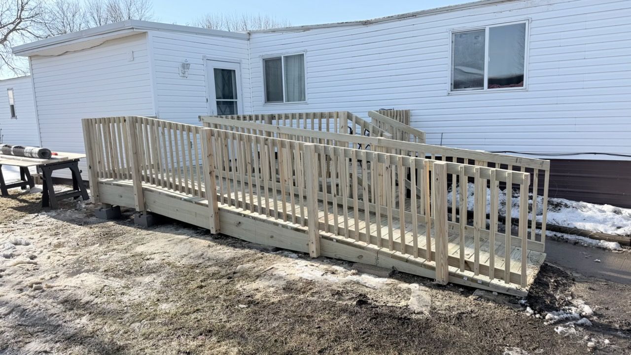 A light-colored wooden wheelchair ramp with railings leading up to the side entrance of a white mobile home.