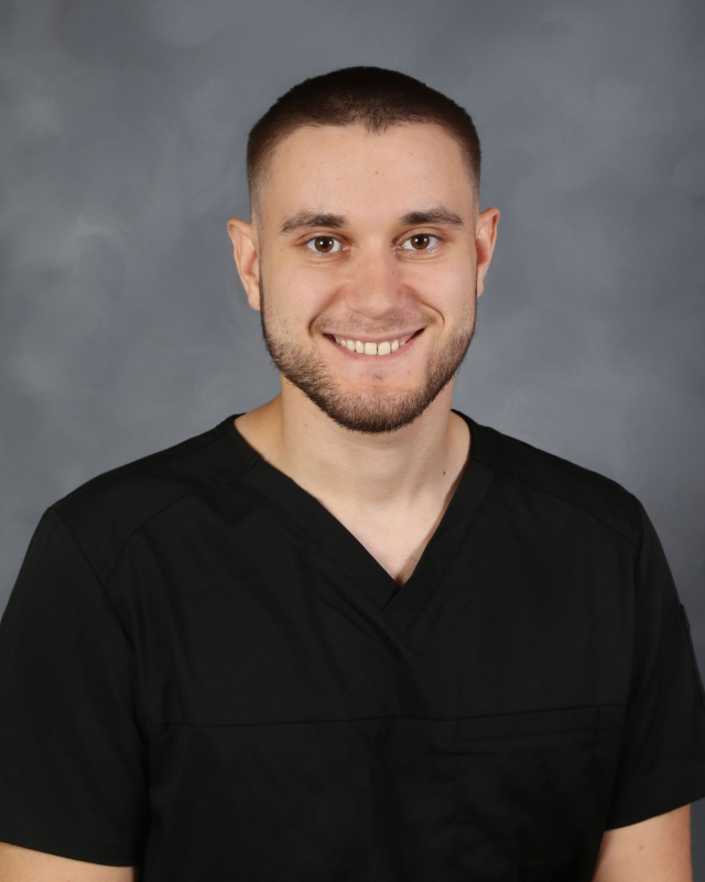 Man in black scrubs smiling, short brown hair, small beard, neutral gray background.