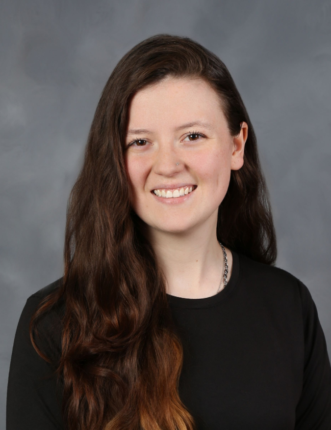 Woman with long brown hair smiles, wearing a black shirt against a gray background.