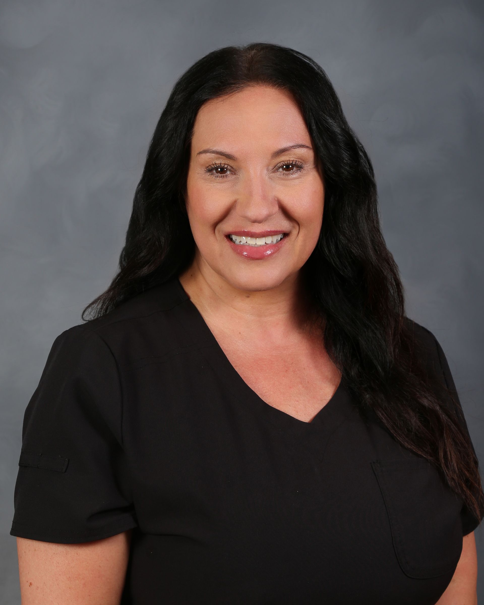 Woman with long dark hair smiling, wearing a black top, against a gray backdrop.