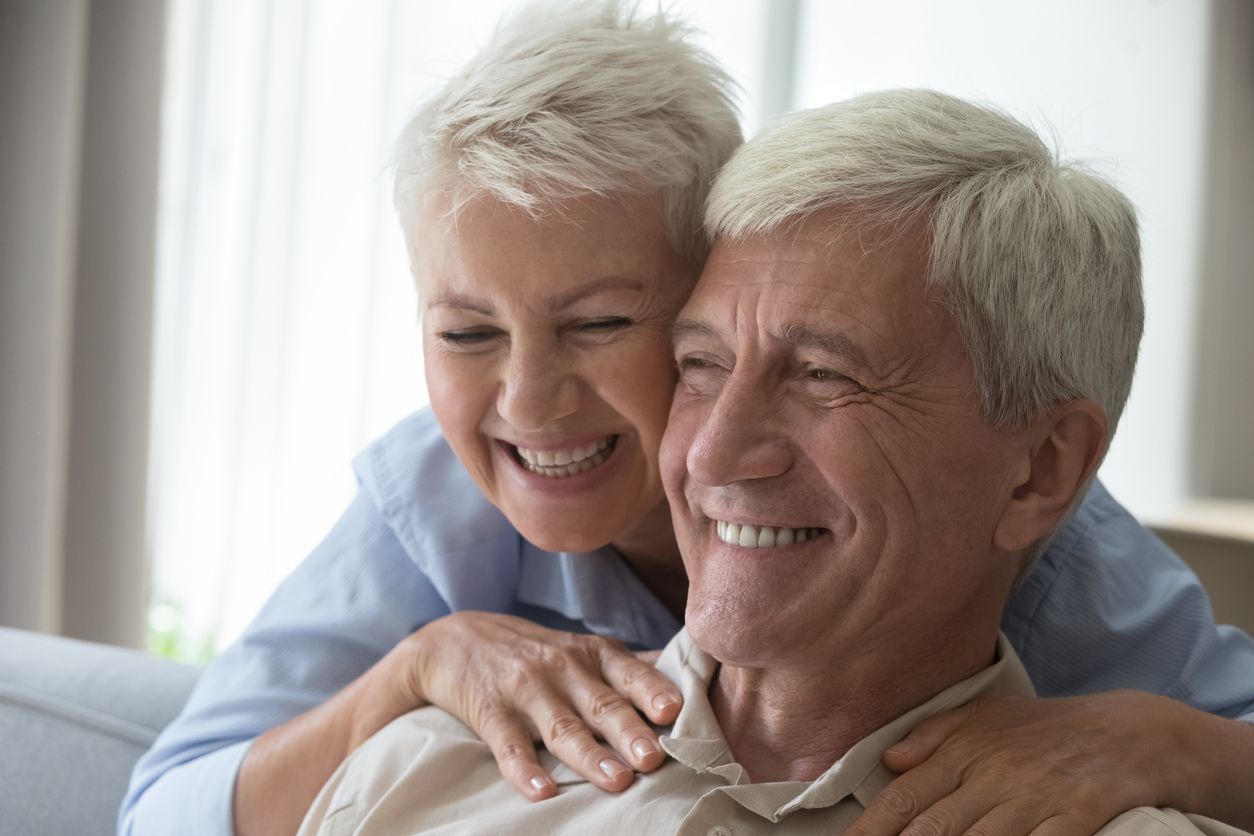 Elderly couple embraces, smiling. Woman hugs man from behind, both with silver hair, indoor.