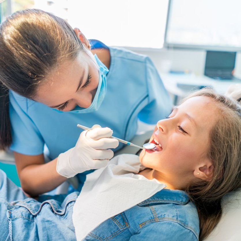 Dentist examining a young girl's teeth in a clinic. Both are focused, the girl's eyes closed.