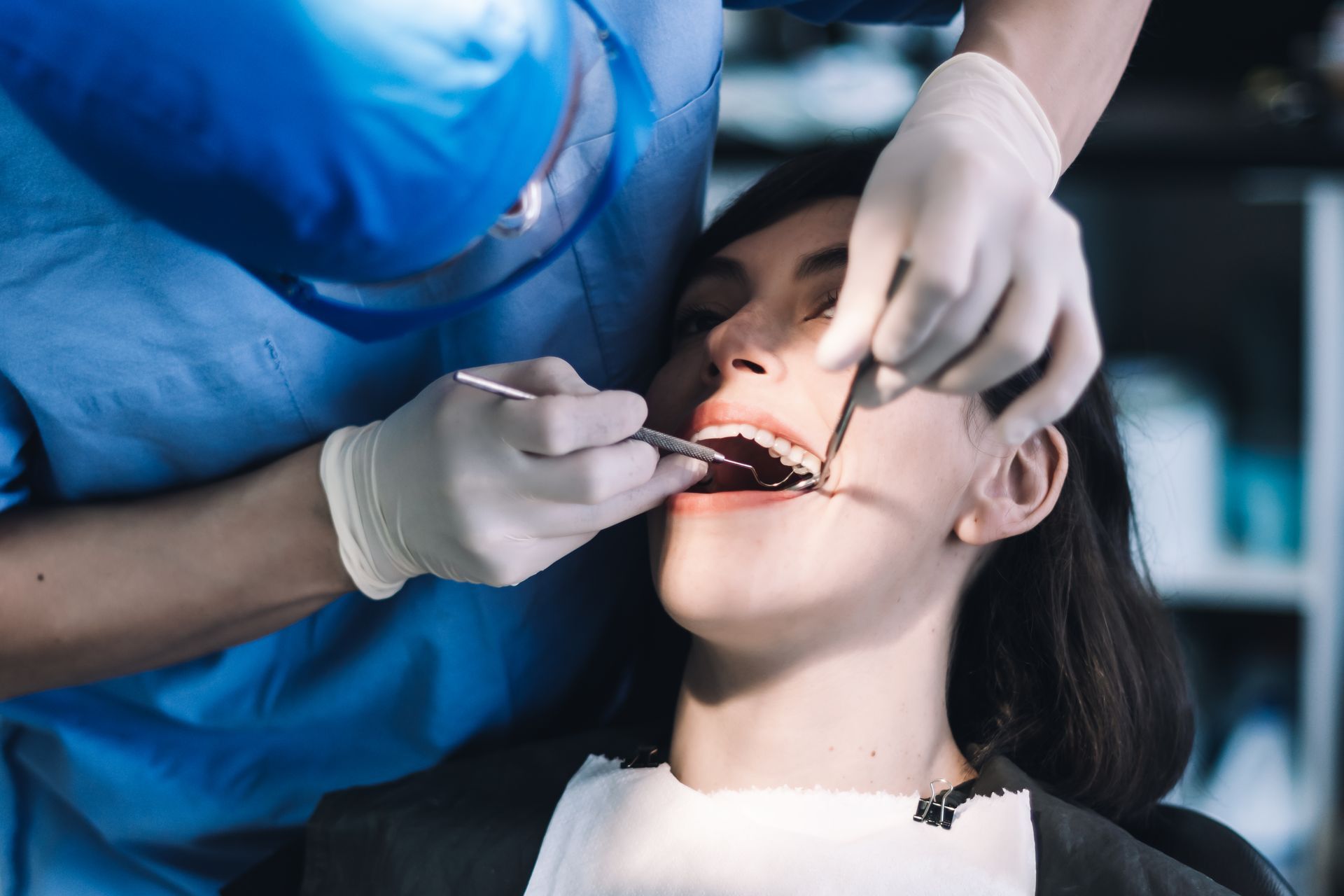 Dentist examining a patient's teeth in a dental clinic. The dentist is wearing gloves and a blue scrub.