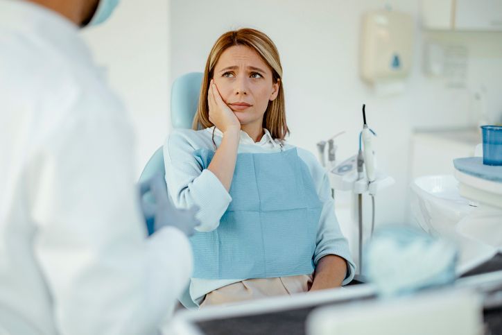 Man in dental chair smiles, looking at teeth in hand mirror. Dentist and assistant in blue scrubs.
