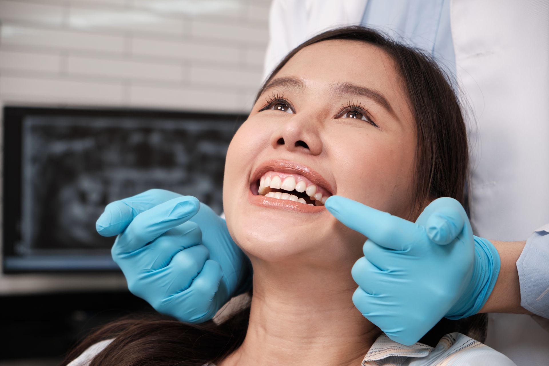 Dentist examining a smiling Asian woman's teeth in a dental office, blue gloves and x-ray visible.