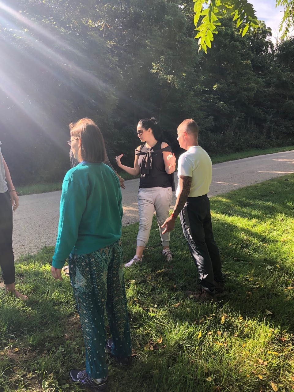 Four people standing outside on a sunny day, conversing on a paved path lined by trees.