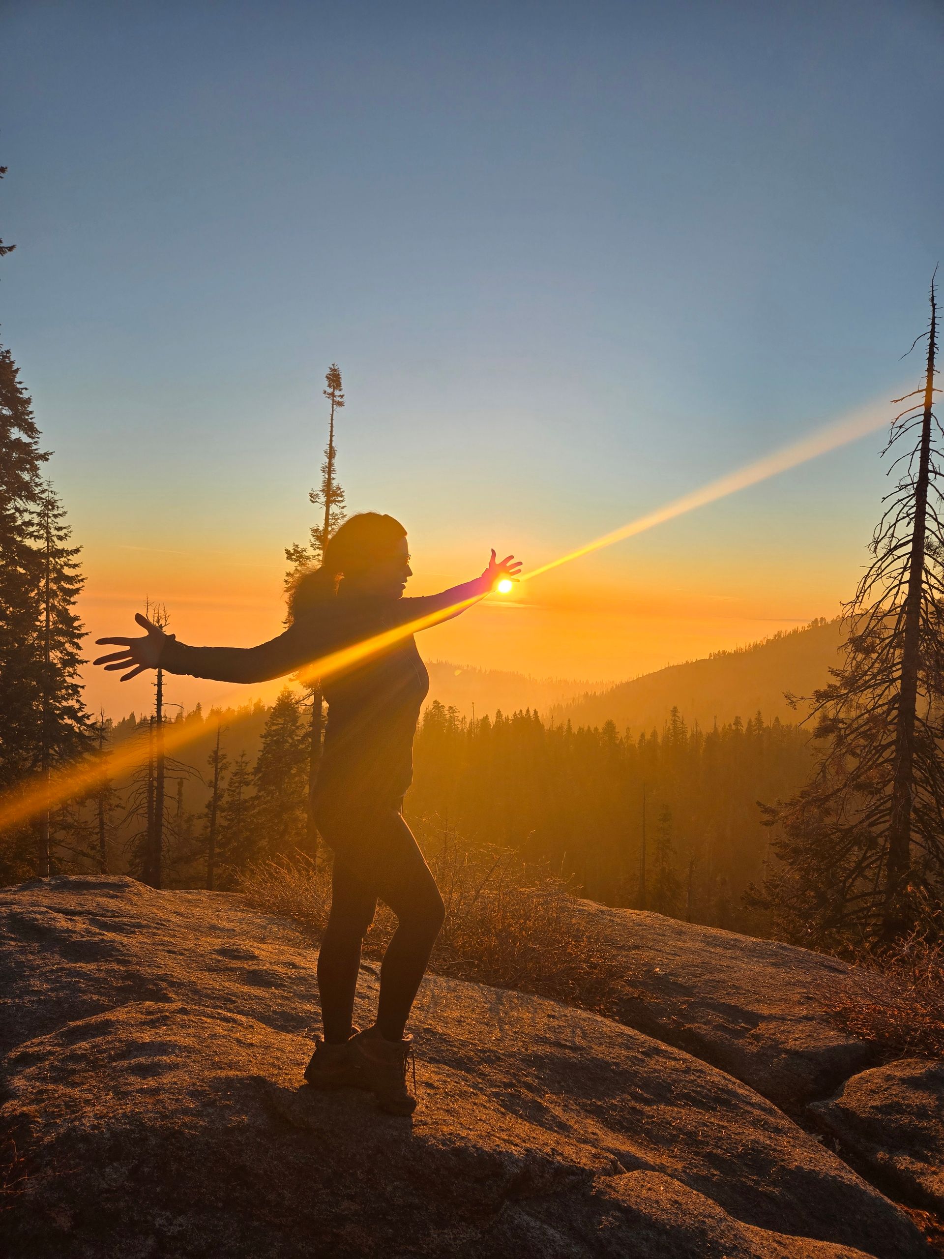 A person stands on a rocky mountain ledge with arms outstretched, backlit by a vibrant, golden sunset over a forest.