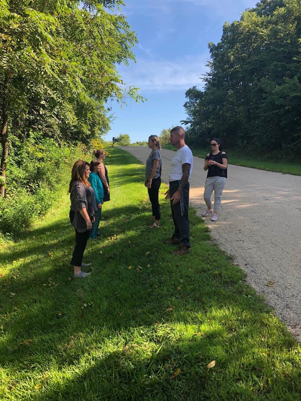 A group of people standing on the edge of a gravel road and grassy area on a sunny day, looking toward the horizon.